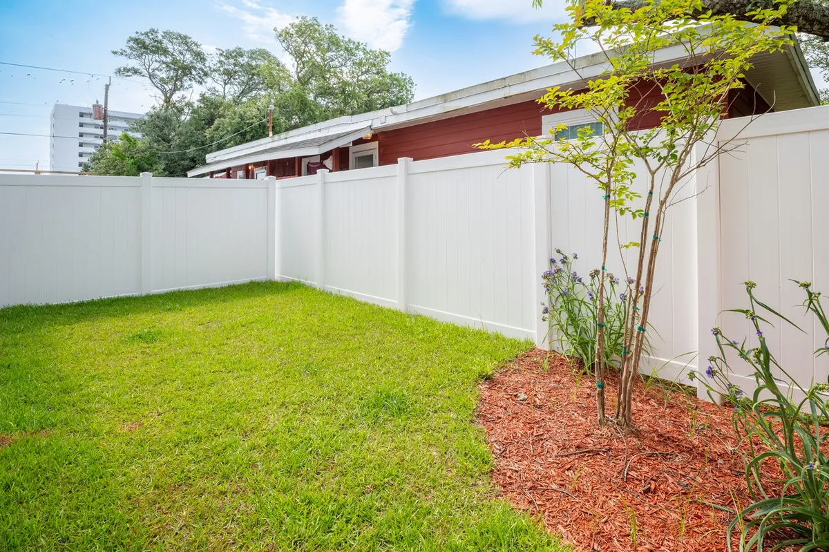 A peaceful exterior scene featuring a vibrant green lawn bordered by a crisp white fence, with a small tree and colorful flower beds adding a touch of nature. The space is bathed in warm sunset light, inviting outdoor relaxation and offering a glimpse into the property's connection with its outdoor environment.