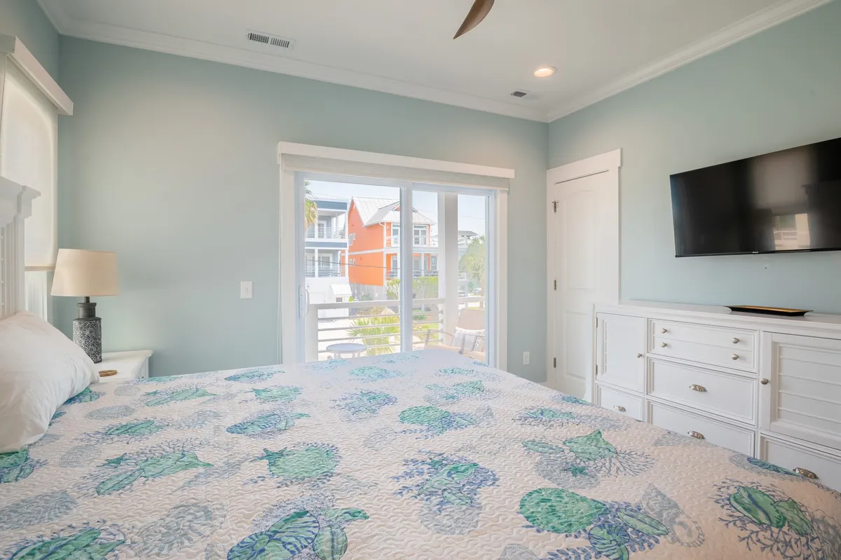 A calming bedroom with a bed topped with a patterned quilt, large windows letting in abundant natural light, a flat-screen TV, and a white dresser. The setting is cozy and modern, ideal for restful nights and peaceful mornings.
