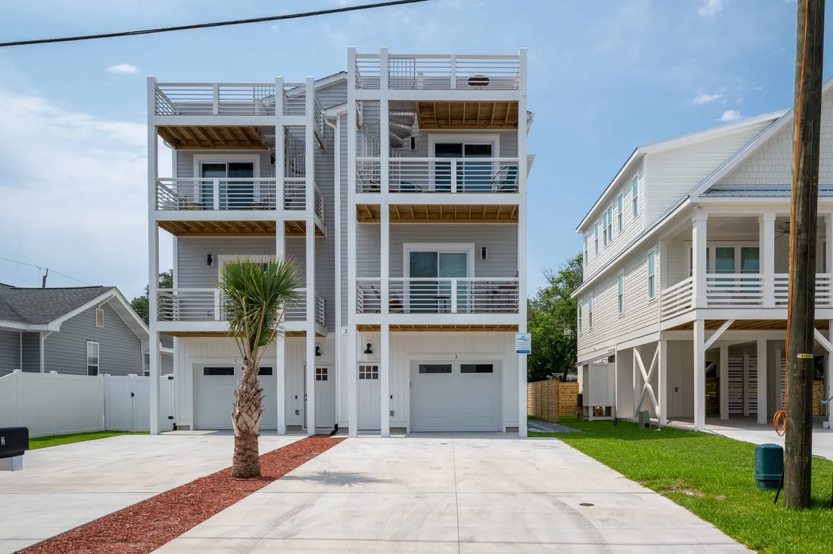 A three-story modern townhouse with multiple balconies, a palm tree in the front yard, and a wide driveway. The building’s clean lines and layered balconies evoke upscale, contemporary coastal living. The balconies suggest outdoor relaxation with sunset or ocean views.