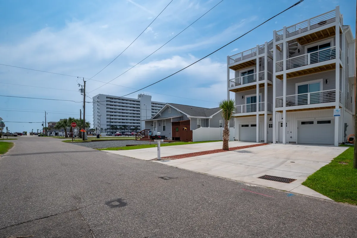 A modern multi-story townhouse stands alongside a classic beach house, with palm trees, a sidewalk, and a street sign setting a relaxed, coastal neighborhood scene. The view hints at walkability and proximity to attractions.