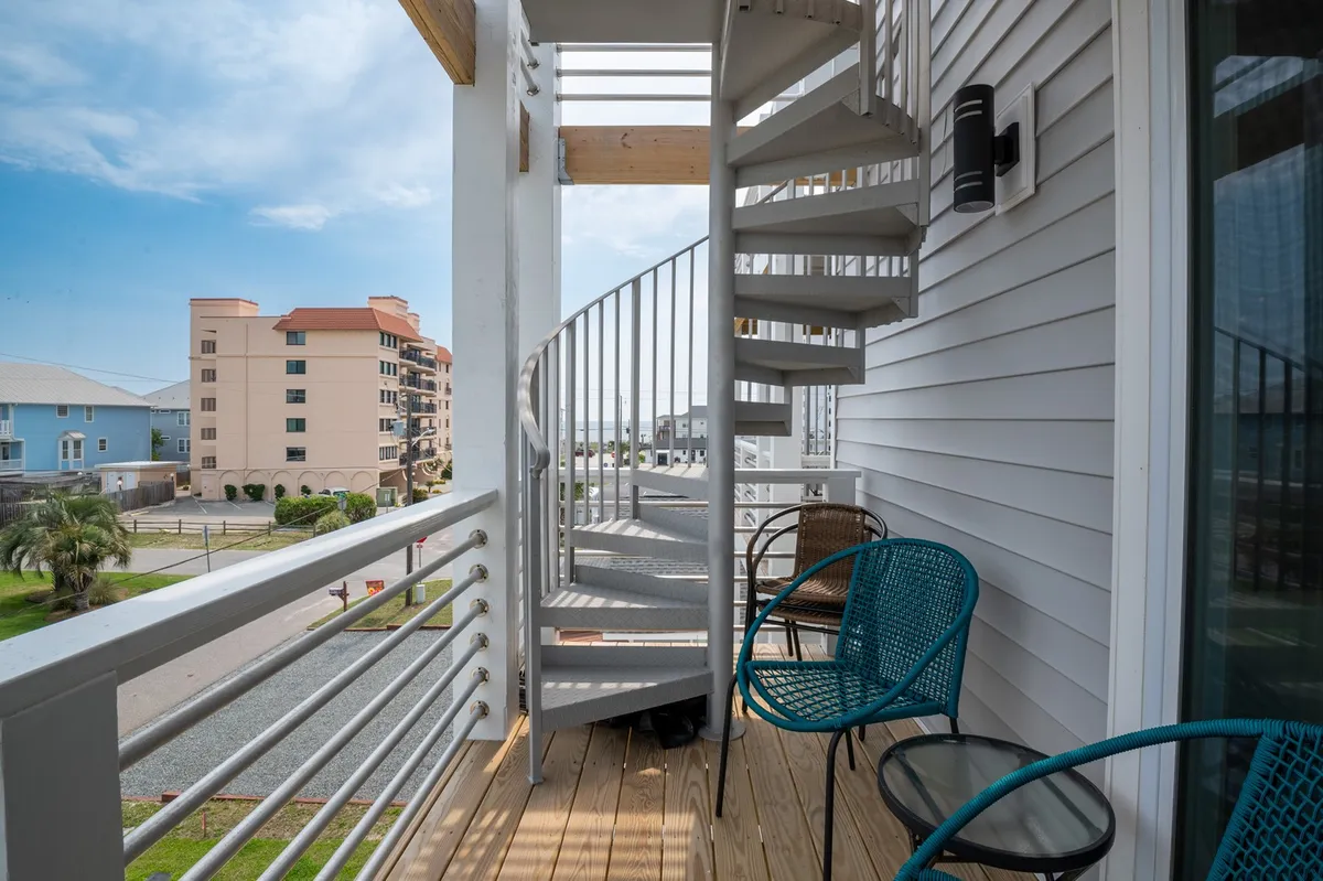 An exterior view of the balcony featuring a spiral staircase, blue chairs, and nearby buildings. The space is clearly designed for relaxation and enjoyment of ocean views, with the spiral staircase hinting at access to the rooftop sun deck.