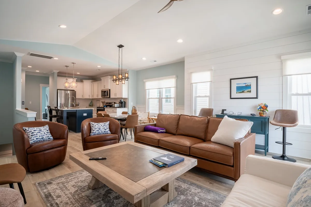 This image features a cozy, open-concept living area with high-end leather sofas, a wooden coffee table, a visible dining area, and stylish light fixtures. The space is bathed in natural light, creating a welcoming environment perfect for socializing or relaxing.