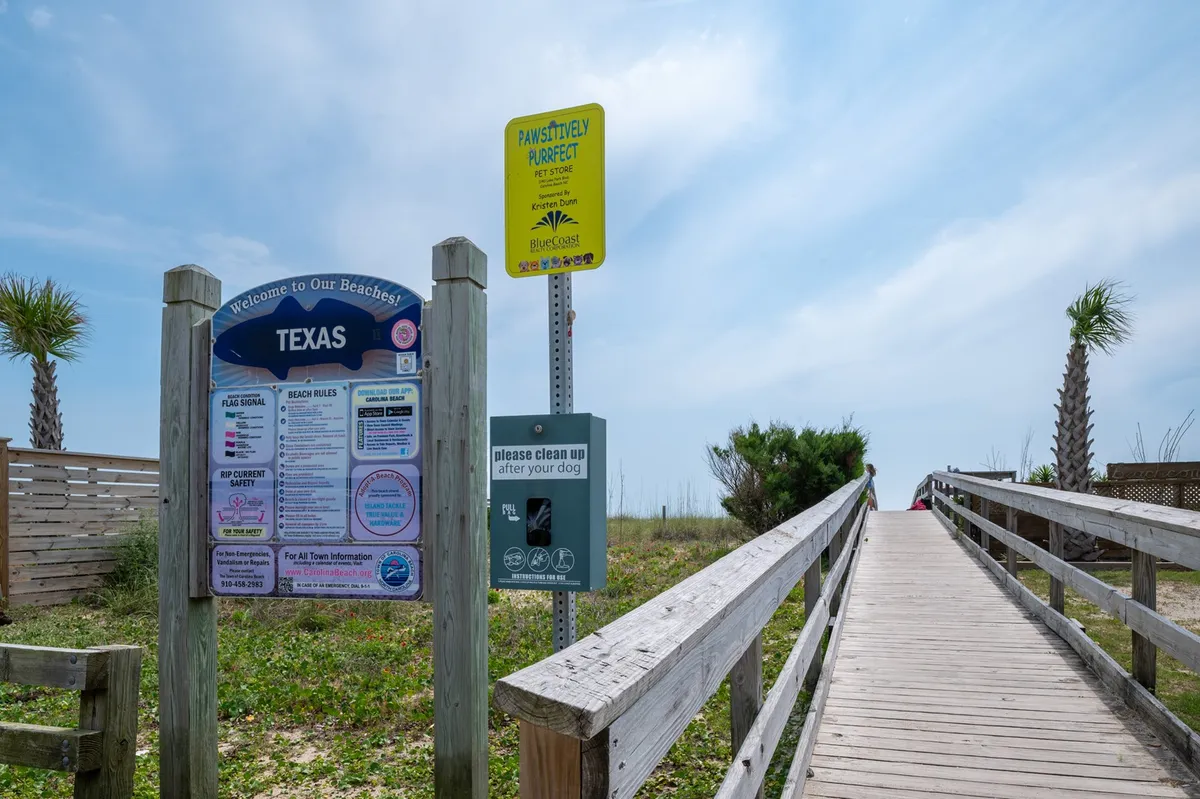 A welcoming pathway with a 'welcome' sign, beach rules, and a dog clean-up station leads directly to the sandy shore. The scene is friendly and approachable, inviting families and groups to step right onto the beach.