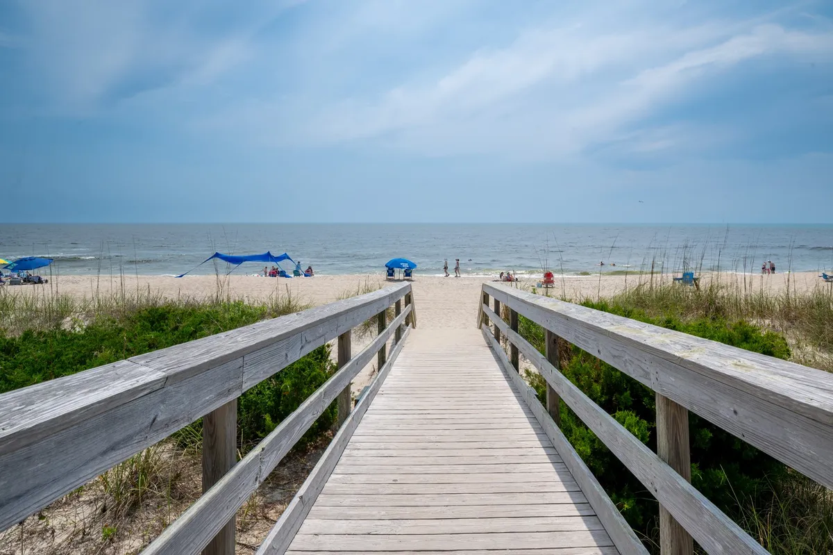 This image features a wooden walkway leading directly to a wide, sandy beach with the sparkling ocean and blue skies beyond. The inviting path draws the eye toward the shoreline, conjuring scenes of family beach days, sunbathing, swimming, and playing in the sand.