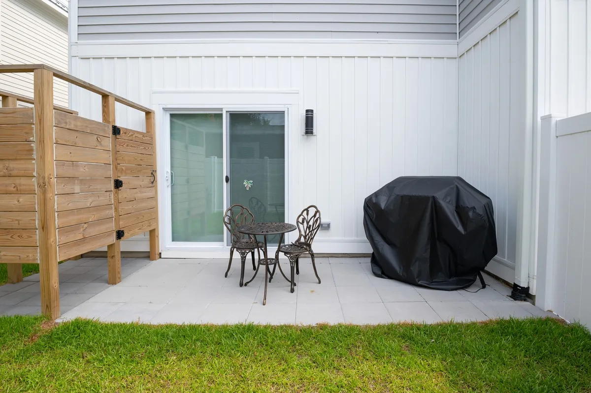 A cozy outdoor space featuring a bistro table and chairs, grill cover, sliding glass door, and a wooden privacy screen—suggestive of relaxed evenings dining al fresco or socializing before exploring Carolina Beach nightlife.