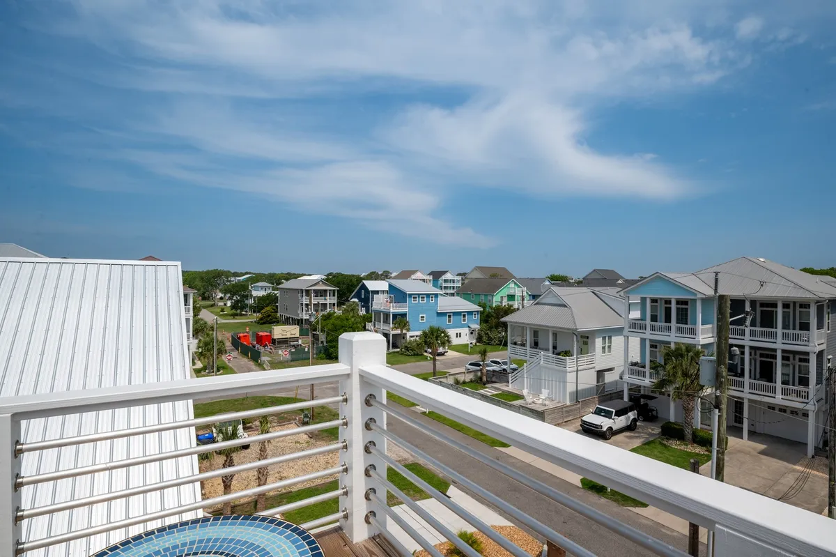 A vibrant balcony view showing colorful houses, a blue sky, and a balcony railing. The environment is perfect for group socialization, morning coffee, or winding down after a night out. The vista hints at the lively, walkable neighborhood and proximity to the boardwalk.