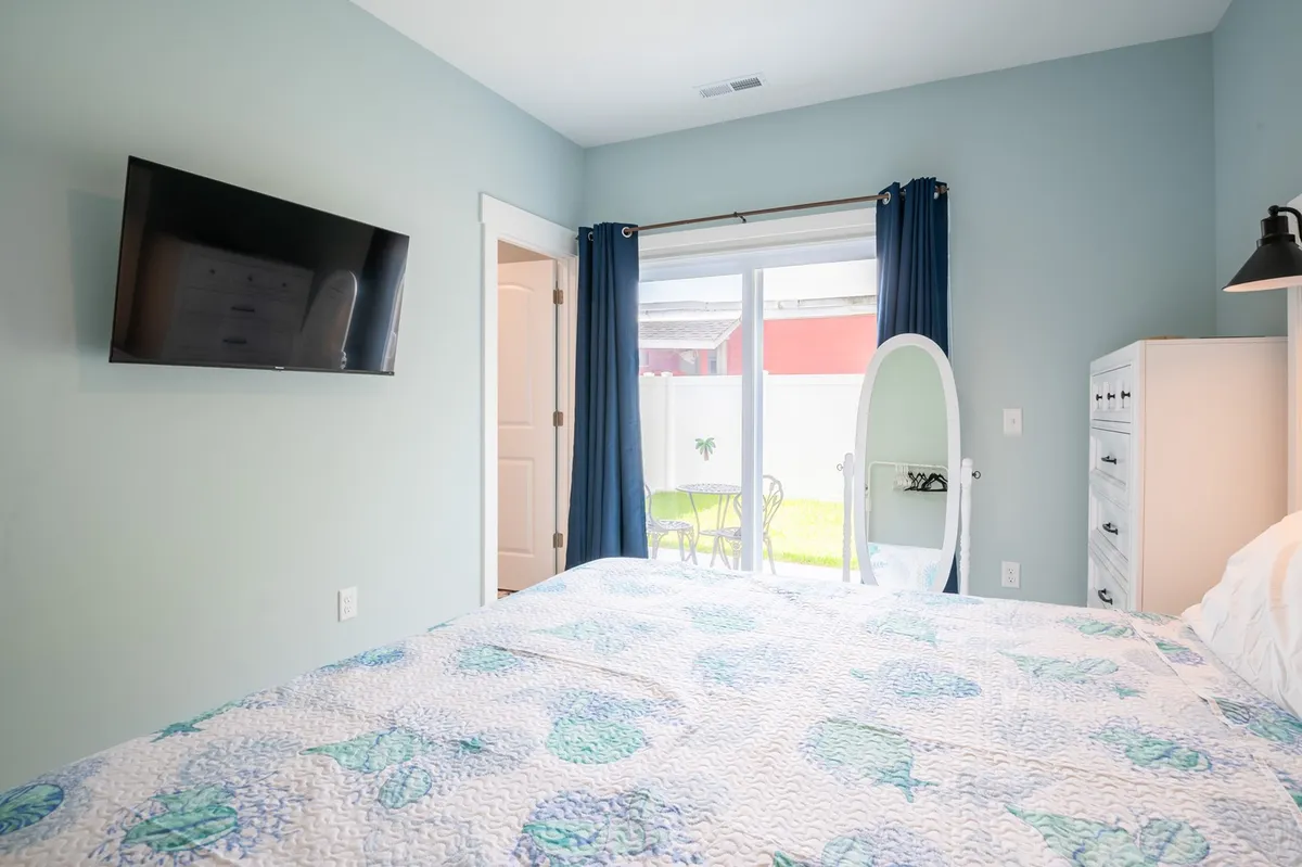 A serene bedroom featuring a bed with a vibrant quilt, a large mirror, wall-mounted TV, and a sliding glass door leading to a private outdoor space. The room is bathed in natural light, emphasizing comfort and privacy.